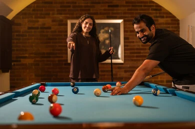 Smiling and playing between partners, successful marital relationships, favorite times between spouses, dialogue and discussion among friends, billiard café, billiard table (snooker), a man and his wife, Arab Saudi Gulf nationals, dressed in casual attire playing billiards, a youth sports hall.