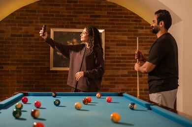 A selfie to document beautiful moments, smiles and play between partners, successful marital relationships, favorite times between couples, dialogue and discussion among friends, a billiards café, a billiards table (snooker), a man and his wife, Arab Saudi Gulf nationals, dressed in casual attire playing billiards.