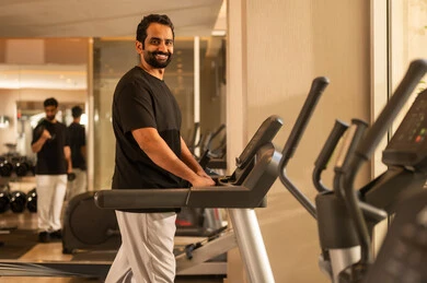Looking at the camera with signs of joy, focusing on health and physical fitness, a youthful activity that helps with physical flexibility. A Saudi man is practicing an exercise to improve body posture and balance. A young Arab Gulf Saudi man wearing sportswear is exercising on a treadmill, using fitness equipment.