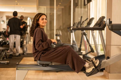 Physical activities to maintain fitness, enjoyable time during exercise, healthy lifestyle, use of modern technological devices, concept of burning fat and losing weight, image of a Saudi Gulf Arab girl wearing sportswear, listening to music while exercising on a stationary bike.
