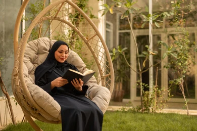 الاستمتاع في مطالعة الكتب والروايات، شابة عربية خليجية سعودية ترتدي عباءة تجلس على الارجوحة تقوم بقراءة كتاب، قضاء وقت ممتع في الهواء الطلق في احدى الشاليهات السياحية