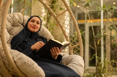الاستمتاع في مطالعة الكتب والروايات، شابة عربية خليجية سعودية ترتدي عباءة تجلس على الارجوحة تقوم بقراءة كتاب، قضاء وقت ممتع في الهواء الطلق في احدى الشاليهات السياحية