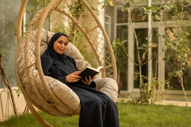 قضاء وقت الفراغ في مطالعة الكتب والروايات، شابة عربية خليجية سعودية ترتدي عباءة تجلس على الارجوحة تقوم بقراءة كتاب، قضاء وقت ممتع في الهواء الطلق في احدى الشاليهات السياحية