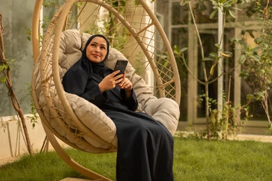 تستخدم امرأة سعودية شابة من الخليج العربي ترتدي العباءة جهازًا تقنيًا حديثًا ومتقدمًا، تجلس على أرجوحة ممسكة بهاتف محمول، تستمتع بوقتها في الهواء الطلق في أحد الشاليهات السياحية، تتواصل مع العائلة والأصدقاء.