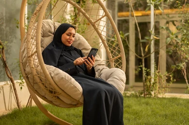 تستخدم امرأة سعودية شابة من الخليج العربي ترتدي العباءة جهازًا تقنيًا حديثًا ومتقدمًا، تجلس على أرجوحة ممسكة بهاتف محمول، تستمتع بوقتها في الهواء الطلق في أحد الشاليهات السياحية، تتواصل مع العائلة والأصدقاء.