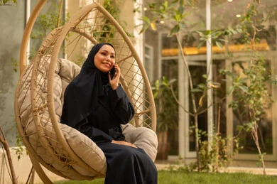 إجراء مكالمة هاتفية عبر الهاتف المحمول، باستخدام جهاز تقني حديث ومتقدم، امرأة سعودية شابة من الخليج العربي ترتدي العباءة جالسة على أرجوحة تحمل هاتفًا محمولًا، تستمتع بوقتها في الهواء الطلق في أحد الشاليهات السياحية، تتواصل مع العائلة والأصدقاء.
