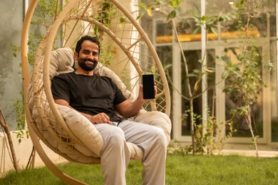 A blank black screen displayed on the mobile phone, chatting with family and friends, using a modern technological device, a young Arab Gulf Saudi man wearing casual attire holding a mobile phone and sitting outdoors, the outdoor setting in one of the chalets, expressions and gestures of happiness and joy.