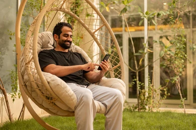 الدردشة مع الاهل والاصدقاء، استخدام جهاز تقني حديث، شاب عربي خليجي سعودي يرتدي الزي الكاجوال يقوم بتصفح مواقع التواصل عبر الهاتف المحمول ويجلس في الهواء الطلق، الجلسة الخارجية في احدى الشاليهات، تعابير وايماءات السعادة والسرور