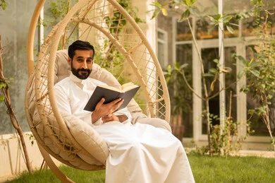 The concept of self-learning and self-cultivation, an Arab Gulf Saudi man wearing traditional attire sitting outdoors reading a book, enjoying time with himself, developing his vocabulary.
