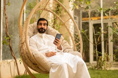 Chatting with family and friends, using modern technology, a young Saudi Arabian Gulf man wearing traditional attire browsing social media on his mobile phone while sitting outdoors, the outdoor setting in one of the chalets, expressions and gestures of happiness and joy.