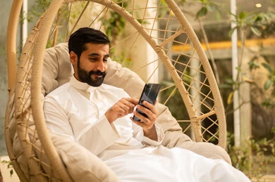 Chatting with family and friends, using modern technology, a young Saudi Arabian Gulf man wearing traditional attire browsing social media on his mobile phone while sitting outdoors, the outdoor setting in one of the chalets, expressions and gestures of happiness and joy. Chatting with family and friends, using modern technology, a young Saudi Arabian Gulf man wearing traditional attire browsing social media on his mobile phone while sitting outdoors, the outdoor setting in one of the chalets, expressions and gestures of happiness and joy.