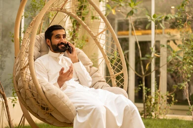 Using a modern technical device, a young Arab Gulf Saudi man wearing traditional attire is making a phone call via a mobile phone while sitting outdoors, in an outdoor setting at one of the chalets, responding to the phone call with expressions of joy.