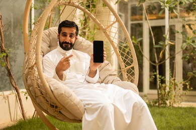 Displaying a blank black screen on the mobile, chatting with family and friends, using a modern technological device, a young Arab Gulf Saudi man wearing traditional attire holding a mobile phone and sitting outdoors, the outdoor setting in one of the chalets, expressions and gestures of happiness and joy.
