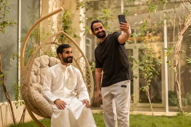 التقاط صورة سيلفي تذكارية باستخدام الهاتف المحمول، تعارف وبناء علاقات اجتماعية، ايماءات السعادة والسرور، شاب عربي خليجي سعودي يرتدي الزي الكاجوال يقضي وقت ممتع مع صديقه في الحديقة، قضاء وقت ممتع في الهواء الطلق في احدى الشاليهات السياحية في المملكة