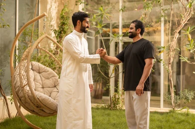 تعارف وبناء علاقات اجتماعية، القاء التحية والسلام، ايماءات السعادة والسرور، شاب عربي خليجي سعودي يرتدي الزي الكاجوال يقوم بمصافحة صديقه في الحديقة، قضاء وقت ممتع في الهواء الطلق في احدى الشاليهات السياحية في المملكة العربية السعودية