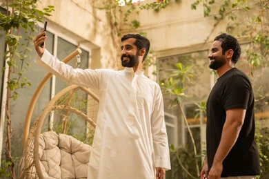 التقاط صورة سيلفي تذكارية باستخدام الهاتف المحمول، تعارف وبناء علاقات اجتماعية، ايماءات السعادة والسرور، شاب عربي خليجي سعودي يرتدي الزي الكاجوال يقضي وقت ممتع مع صديقه في الحديقة، قضاء وقت ممتع في الهواء الطلق في احدى الشاليهات السياحية في المملكة