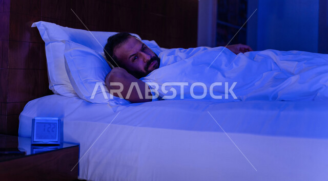 رجل عربي خليجي سعودي مستلقي على فراشه وامامه ساعة تنبيه، وسادة بيضاء، الأرق الليلي، القلق والإكتئاب، التفكير ومحاولة النوم والراحة