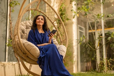 تستخدم امرأة سعودية شابة من الخليج العربي ترتدي العباءة جهازًا تقنيًا حديثًا ومتقدمًا، تجلس على أرجوحة ممسكة بهاتف محمول، تستمتع بوقتها في الهواء الطلق في أحد الشاليهات السياحية، تتواصل مع العائلة والأصدقاء.