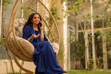 Making a phone call via mobile, using a modern and advanced technical device, a young Arab Gulf Saudi woman wearing an abaya sitting on a swing holding a mobile phone, enjoying time outdoors in one of the tourist chalets, communicating with family and friends.
