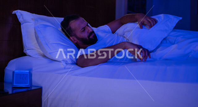رجل عربي خليجي سعودي مستلقي على فراشه وامامه ساعة تنبيه، وسادة بيضاء، الأرق الليلي، القلق والإكتئاب، التفكير ومحاولة النوم والراحة