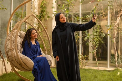 Taking a commemorative selfie using a mobile phone, using a modern and advanced technical device, a young Arab Gulf Saudi woman wearing an abaya enjoying time with her friend in the park, having a good time outdoors in one of the tourist chalets.