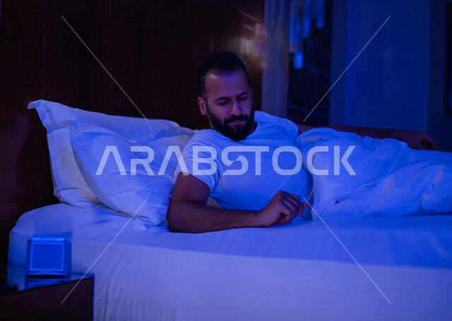 A Saudi Arabian Gulf man lying on his bed with an alarm clock in front of him, a white pillow, night insomnia, anxiety and depression, thinking and trying to sleep and rest