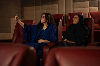 الاستمتاع بمشاهدة الأفلام داخل السينما،متابعة الفيلم المفضل، ،تعابير الاندماج والتفاعل ،  امرأة عربية خليجية سعودية محجبة ترتدي عباءه مع صديقتها ، استخدام الهاتف لتصوير سيلفي للذكرى