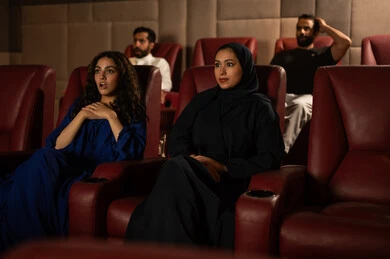 Enjoying watching movies in the cinema, following the favorite film, expressions of engagement and interaction, an Arab Gulf Saudi woman wearing a hijab and an abaya with her friend, fun and entertainment by spending enjoyable times.