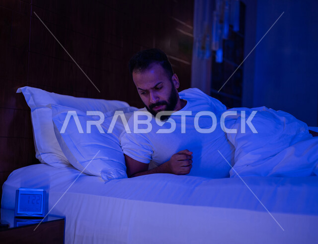 رجل عربي خليجي سعودي مستلقي على فراشه وامامه ساعة تنبيه، وسادة بيضاء، الأرق الليلي، القلق والإكتئاب، التفكير ومحاولة النوم والراحة