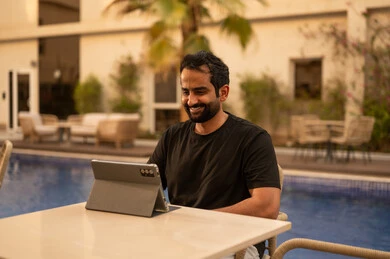 تصفح مواقع التواصل الاجتماعي عبر الجهاز اللوحي، الدردشة مع الاهل والاصدقاء، استخدام جهاز تقني حديث، شاب عربي خليجي سعودي يرتدي الزي الكاجوال يستخدم التابليت المحمول ويجلس في الهواء الطلق، الجلسة الخارجية في احدى الشاليهات، تعابير وايماءات السعادة والسرور