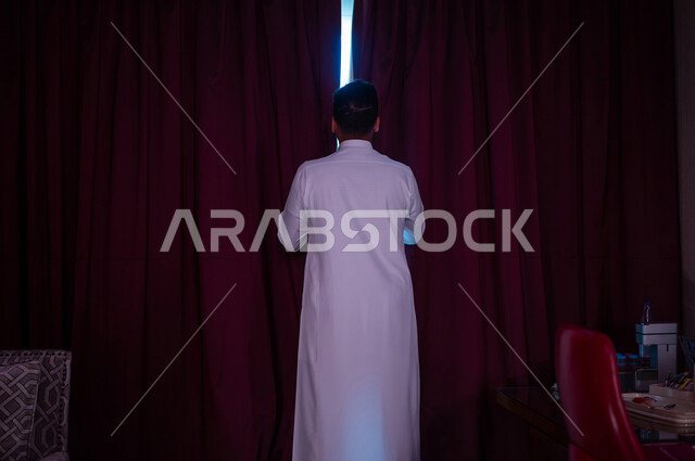 رجل عربي خليجي سعودي يقوم بفتح ستارة النافذة، غرفة المكتب، النظر الى الخارج من خلال النافذة، إشراقة يوم جديد، مفهوم الهمة والنشاط