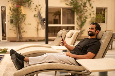 Browsing social media on a mobile phone, chatting with family and friends, using a modern technological device, a young Arab Gulf Saudi man wearing casual attire holding a mobile phone sitting outdoors, the outdoor setting in one of the chalets, expressions and gestures of happiness and joy.