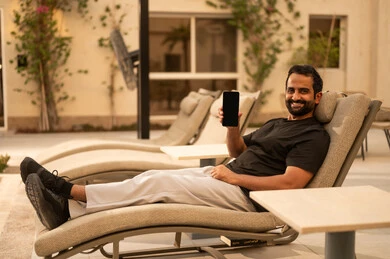 عرض شاشة سوداء فارغة عبر الجوال، الدردشة مع الاهل والاصدقاء، استخدام جهاز تقني حديث، شاب عربي خليجي سعودي يرتدي الزي الكاجوال يمسك بيده الهاتف المحمول ويجلس في الهواء الطلق، الجلسة الخارجية في احدى الشاليهات، تعابير وايماءات السعادة والسرور