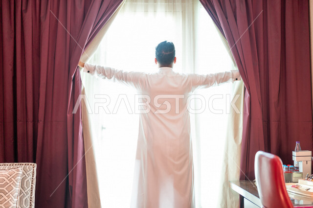 رجل عربي خليجي سعودي يقوم بفتح ستارة النافذة، غرفة المكتب، النظر الى الخارج من خلال النافذة، إشراقة يوم جديد، مفهوم الهمة والنشاط
