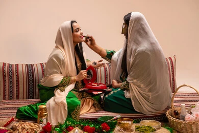 الاحتفال بليلة العمر، منتجات ومساحيق التجميل، العناية والاهتمام بالأناقة والجمال، مفهوم التزين والاطلالة المتألقة، سيدة عربية خليجية اماراتية تقوم بوضع أحمر الشفاه للعروسة، إيماءات السعادة والفرح