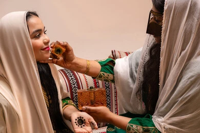 Unique perfumes, joy and happiness on the wedding day, the concept of elegance and attention to appearance, an Emirati Gulf Arab woman wearing a burqa and a white hijab holding a perfume bottle applying fragrance to a bride dressed in traditional attire, wedding ceremonies and celebrations in the UAE.