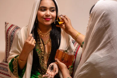 Elegant and exceptional scents, joy and happiness on the wedding day, the concept of elegance and attention to appearance, an Emirati Gulf Arab woman wearing a burqa and a white hijab holding a perfume bottle applying fragrance to a bride dressed in traditional attire, wedding ceremonies and celebrations in the UAE.