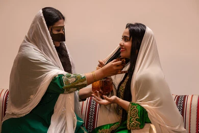 الفرح و السرورفي يوم الزواج، عطور ذات روائح فاخرة وفريدة، مفهوم الأناقة والاهتمام بالمظهر الخارجي، سيدة عربية اماراتية خليجية ترتدي البرقع وحجاب ابيض تحمل زجاجة عطرية تقوم بوضع العطر لعروس ترتدي الزي التراثي، مراسم الأعراس والاحتفالات في الامارات