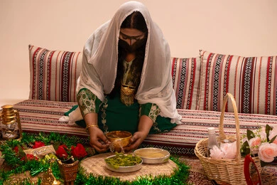 العادات والطقوس الإماراتية في حفلات الزواج، نقوش وزخارف تراثية من مواد طبيعيه، مهنة تقليدية شعبية نسائية، التجمل والتزيين في يوم الزفاف، إمرأة عربية خليجية إماراتية ترتدي البرقع وحجاب ابيض تقوم بتحضير خلطة الحناء