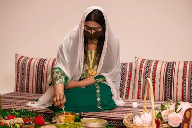 عادات متوارثة تعكس التراث والثقافة المحلية في المناسبات السعيدة ، نقوش وزخارف تراثية من مواد طبيعيه، مهنة تقليدية شعبية نسائية، التجمل والتزيين في يوم الزفاف، إمرأة عربية خليجية إماراتية ترتدي البرقع وحجاب ابيض تقوم بتحضير خلطة الحناء