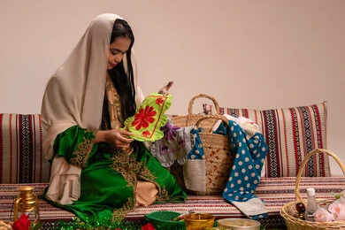 Dressing in traditional folk clothing during occasions and celebrations is a local national product, featuring traditional Emirati heritage and folk art. An Arab Gulf Emirati woman wearing an embroidered abaya holds a piece of fabric made with traditional folk hand embroidery and looks at it.