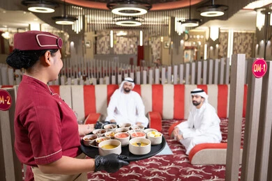 نادله ترتدي زي اليونيفورم تقوم بتقديم الطلبات، الكرم والضيافة عند العرب،قضاء أوقات ممتعة مع الأصدقاء ، رجلان عربيان خليجيان اماراتيان يرتديان الكندورة والغترة يتشاركان الطعام