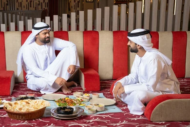Enjoying lunch outdoors, having a good time at the restaurant, restaurants and cafes in the UAE, two Emirati Gulf Arab men wearing kandura and ghutrah, sitting in a modern gathering and sharing food, gestures of happiness and joy.