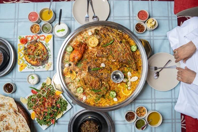 حسن الكرم والضيافة , مطاعم و كافيهات الامارات, صورة مقربة ليد رجلان عربيان خليجيان إماراتيان يرتديان الكندورة والغترة، يجلسان على مائدة مليئة بالمأكولات الشرقية اللذيذة ويتشاركان الطعام, إيماءات السعادة و السرور