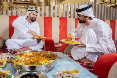 قضاء أوقات ممتعة في المطعم, الاستمتاع بالغداء في الخارج,مطاعم و كافيهات الامارات, رجلان عربيان خليجيان إماراتيان يرتديان الكندورة والغترة، يجلسان في مجلس عصري ويتشاركان الطعام, إيماءات السعادة و السرور
