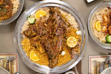 The generosity of hospitality and the authentic Gulf flavor, the tastes of Arab heritage in feasts and occasions, a close-up image of a luxurious traditional dish containing lamb cooked in the mandi style, served over basmati rice seasoned with authentic Arabic spices, garnished with slices of lemon, orange, and fresh vegetables.