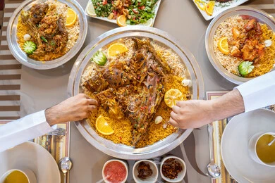 The goodness of generosity and hospitality, restaurants and cafes in the UAE, a close-up image of the hands of two Emirati Gulf Arab men wearing the kandura and ghutrah, sitting at a table filled with delicious Eastern dishes and sharing food, gestures of happiness and joy.
