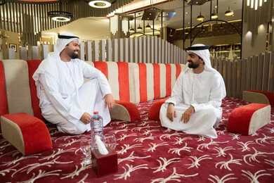 تبادل الأحاديث الودية والابتسامات، رجلان عربيان خليجيان إماراتيان يرتديان الكندورة والغترة، يجلسان في مجلس عصري بأجواء راقية وحديثة ،ايماءات كرم الضيافة وروح التواصل الاجتماعي في المجالس الإماراتية، تصميم داخلي أنيق بإضاءة دافئة ولمسات فخمة ،مفهوم الاصالة والمعاصرة.