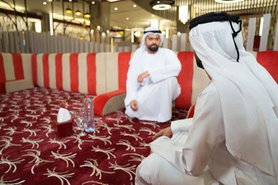 مفهوم الاناقة والفخامة ،رجلان عربيان خليجيان إماراتيان يرتديان الكندورة والغترة، يجلسان في مجلس عصري بأجواء راقية وحديثة، يتبادلان الأحاديث الودية والابتسامات،ايماءات كرم الضيافة وروح التواصل الاجتماعي في المجالس الإماراتية، تصميم داخلي أنيق بإضاءة دافئة ولمسات فخمة ،مفهوم الاصالة والمعاصرة.