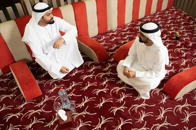 مفهوم الاصالة والمعاصرة، رجلان عربيان خليجيان إماراتيان يرتديان الكندورة والغترة، يجلسان في مجلس عصري بأجواء راقية وحديثة، يتبادلان الأحاديث الودية والابتسامات،ايماءات كرم الضيافة وروح التواصل الاجتماعي في المجالس الإماراتية، تصميم داخلي أنيق بإضاءة دافئة ولمسات فخمة